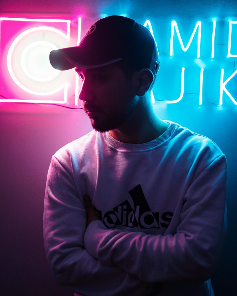 Man in a cap and sweatshirt silhouetted against pink and blue neon lights.